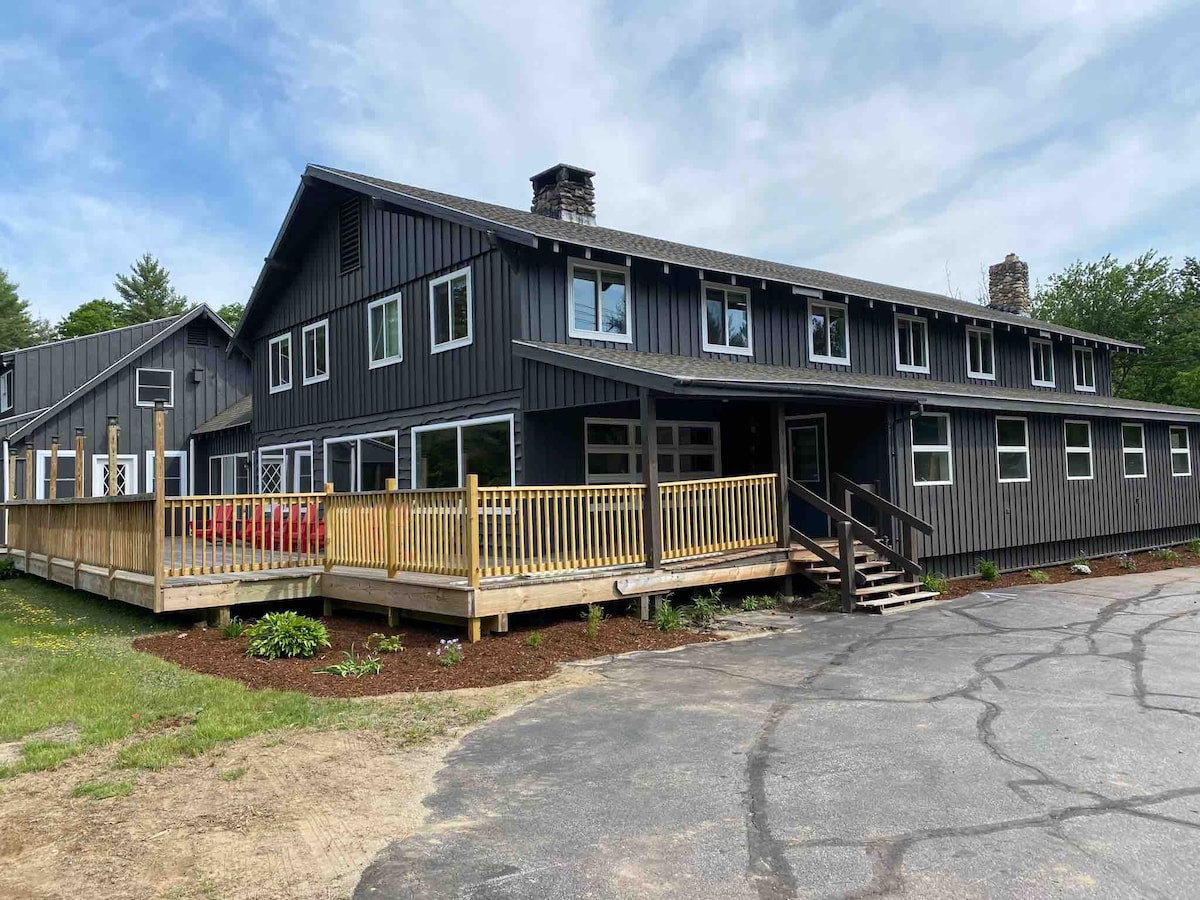 The exterior of The Meeting Haus showcases a modern structure with dark wood siding and multiple large windows. A wooden deck with railings extends from the front, providing access to outdoor seating. A paved parking area is visible, surrounded by green landscaping and trees.