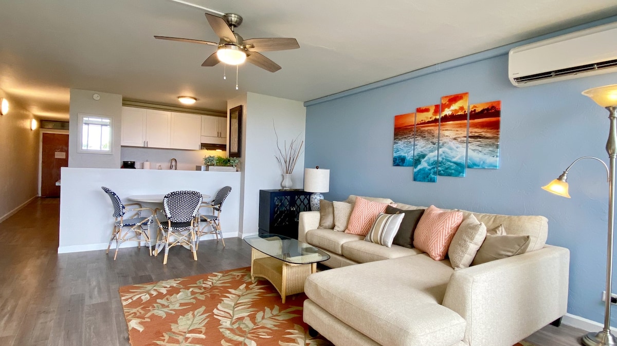 The living area features a comfortable couch with decorative pillows, a glass coffee table, and a patterned area rug. Light-colored walls are complemented by ocean-themed art. A dining space is visible in the background with a modern table and woven chairs. Natural light fills the room.