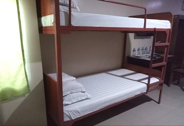 A clean room is presented with sturdy double deck beds. Each bed is neatly made with fresh linens, and bright natural light filters through a nearby window, partially covered by light curtains. A small area for personal items is visible in the background.