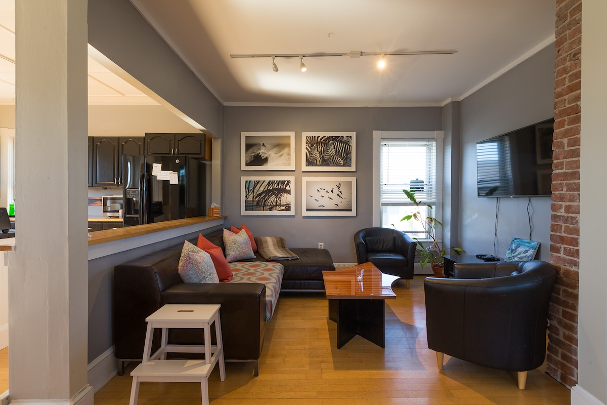 The living area displays contemporary furniture arranged around a coffee table. A cozy couch with patterned pillows faces a flat-screen TV mounted on the wall. Art pieces hang on the opposite wall, and natural light enters through a window, enhancing the inviting space.