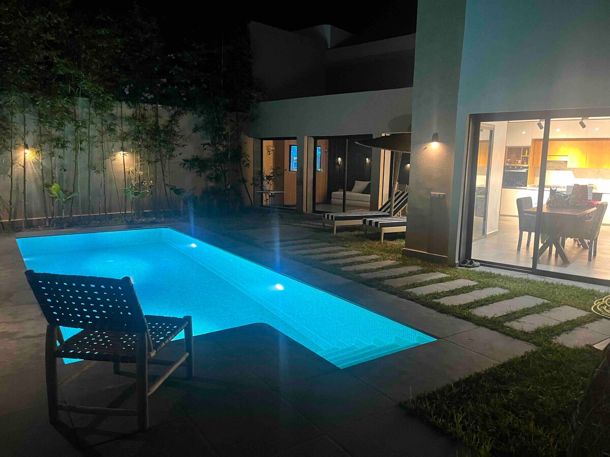 A serene evening view of a well-lit swimming pool, featuring soft blue illumination. Comfortable lounge chairs are positioned nearby, with a grassy area and stepping stones leading toward the indoor living space visible through expansive glass doors.