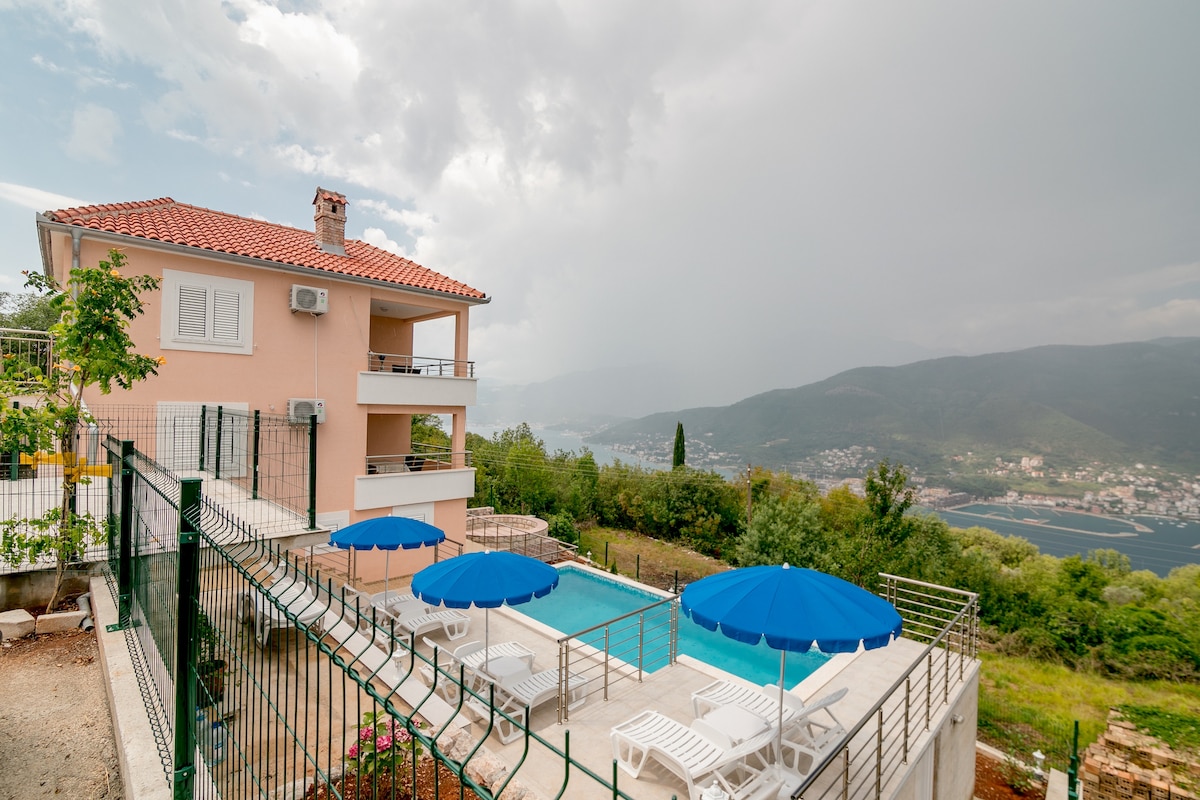 A multi-level house is positioned on a hillside, featuring a private pool surrounded by several sunbeds under large blue umbrellas. Lush greenery frames the property, and scenic views of the valley and distant mountains are visible, creating a serene outdoor setting.