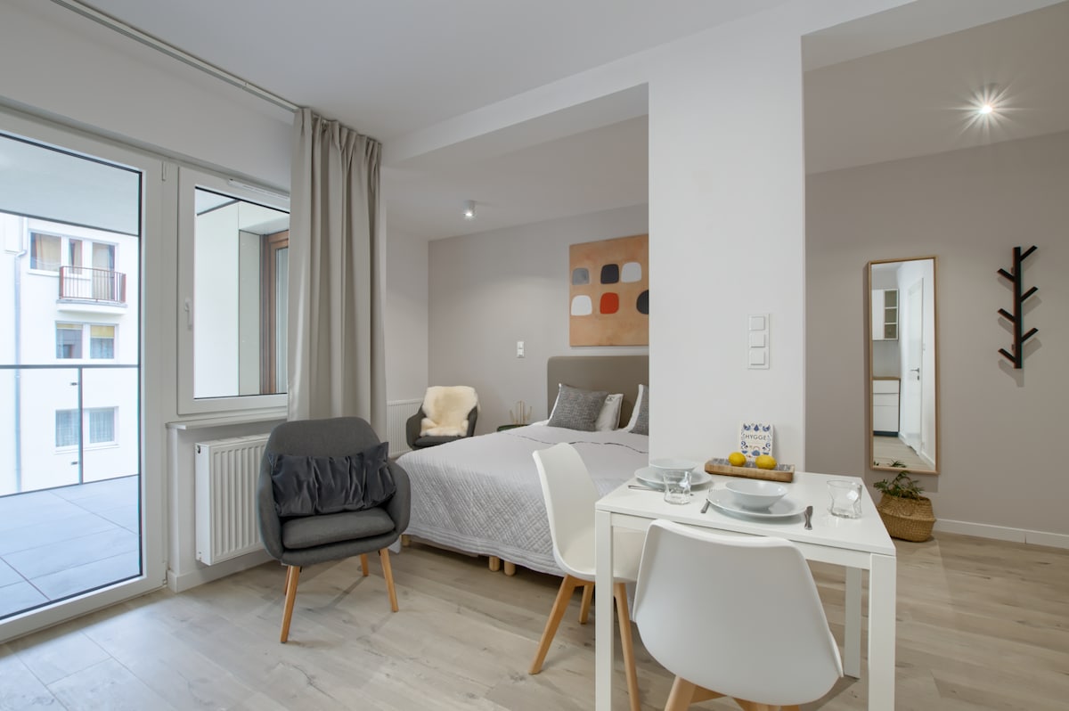 A modern living area features a spacious bed with neutral bedding and a decorative wall art piece above. A small dining table is set for two, positioned near large windows that allow natural light to fill the room. A cozy chair and a mirror add functionality.