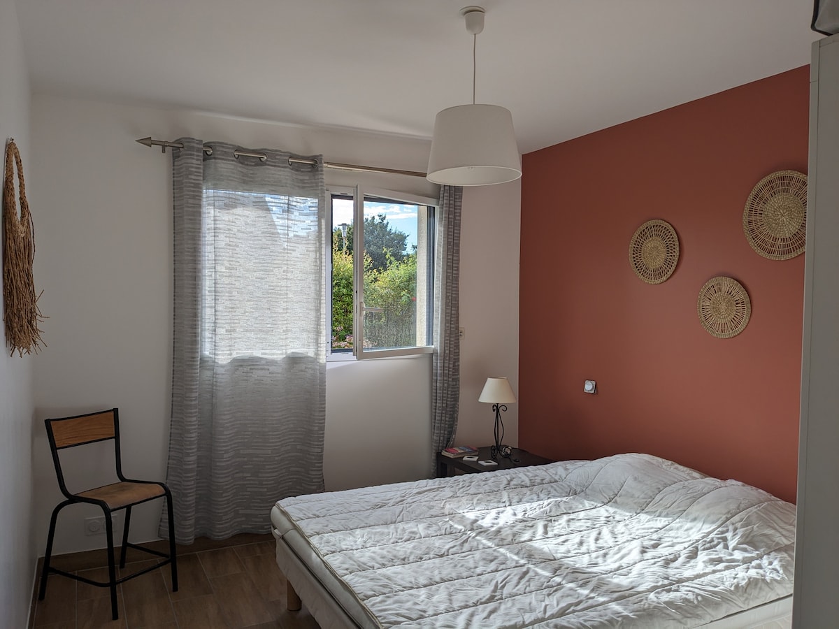 A cozy guest room is presented with a spacious bed featuring a simple white coverlet. Natural light fills the room through a large window, while a chair is positioned in the corner. Decorative wall hangings add a touch of character to the warm-colored accent wall.