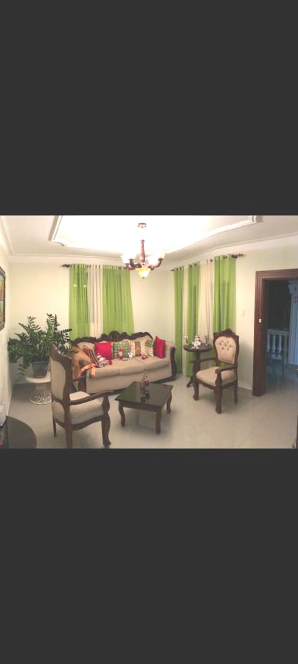 A well-lit living area is presented with elegant wooden furniture, including a cozy sofa adorned with colorful cushions. A pair of armchairs complement the space, which features decorative plants and soft green curtains framing the windows. Natural light enhances the inviting atmosphere.
