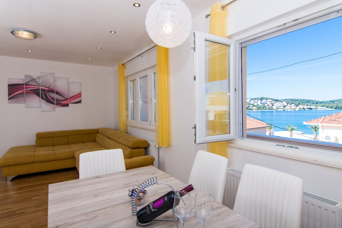 A modern living area features a large yellow sectional sofa and a dining table set for four. Bright yellow curtains frame the windows, which offer a view of the sea and surrounding landscape. Natural light highlights the wood flooring and simple artwork on the wall.