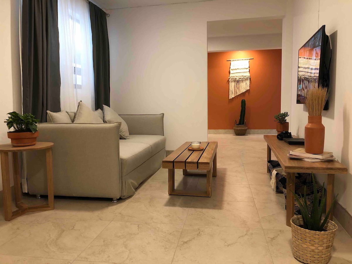 A spacious living area is presented with a soft grey sofa adorned with cushions. A wooden coffee table sits in front, and a flat-screen TV is mounted on the wall opposite. Decorative plants and wall art contribute to a calming, modern aesthetic.