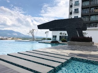 An expansive infinity pool is depicted, featuring a modern structure resembling a sculpture nearby. Smooth, wide steps lead from the pool edge to the building. The backdrop presents a serene landscape with distant mountains under a partly cloudy sky.