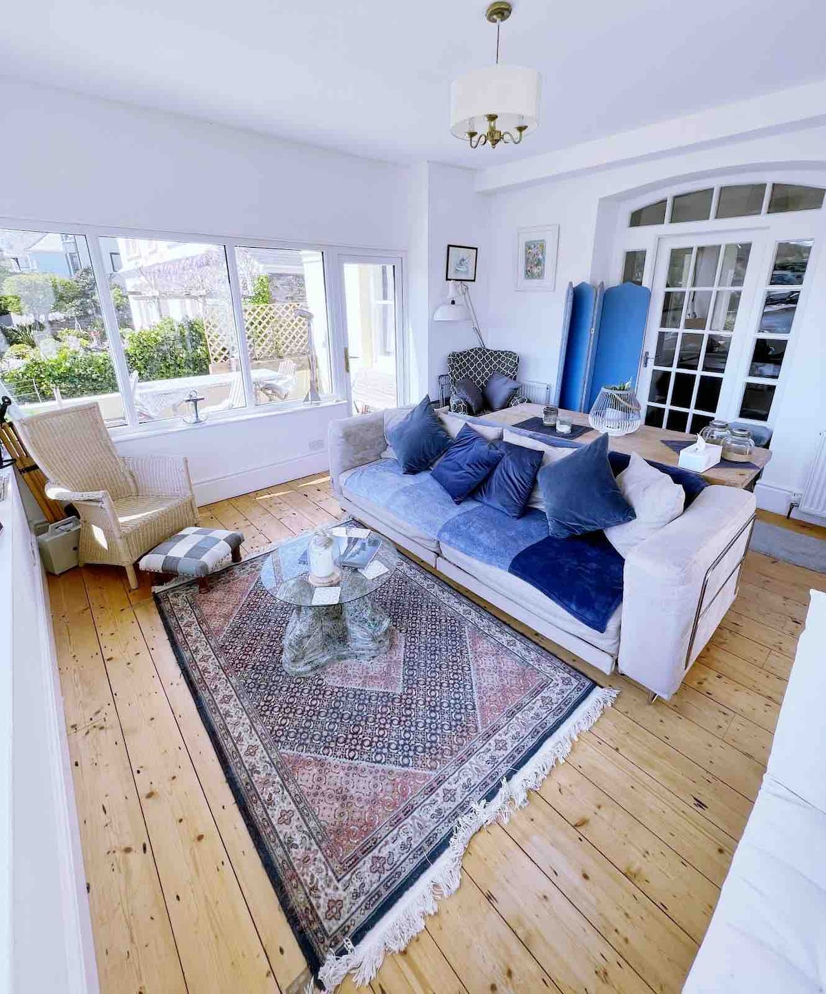 A living area is observed with soft seating arranged around a glass coffee table. A patterned rug covers the wooden floor, while large windows allow natural light to fill the space. Decor elements include framed artwork and a small side table.