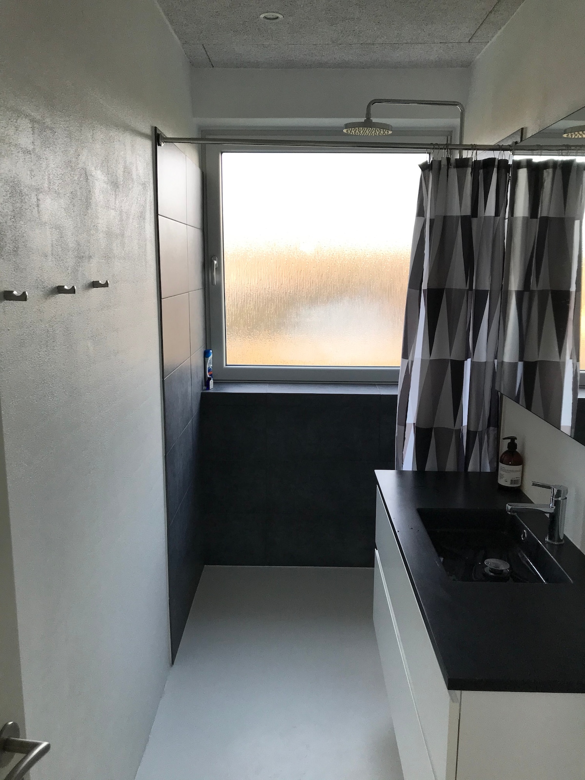 A modern bathroom features a sleek dark countertop alongside a basin and faucet. Neutral-colored walls complement the geometric-patterned shower curtain. A large window allows natural light, with droplets collecting on the glass, creating a serene backdrop. Hooks are visible on the wall for hanging items.