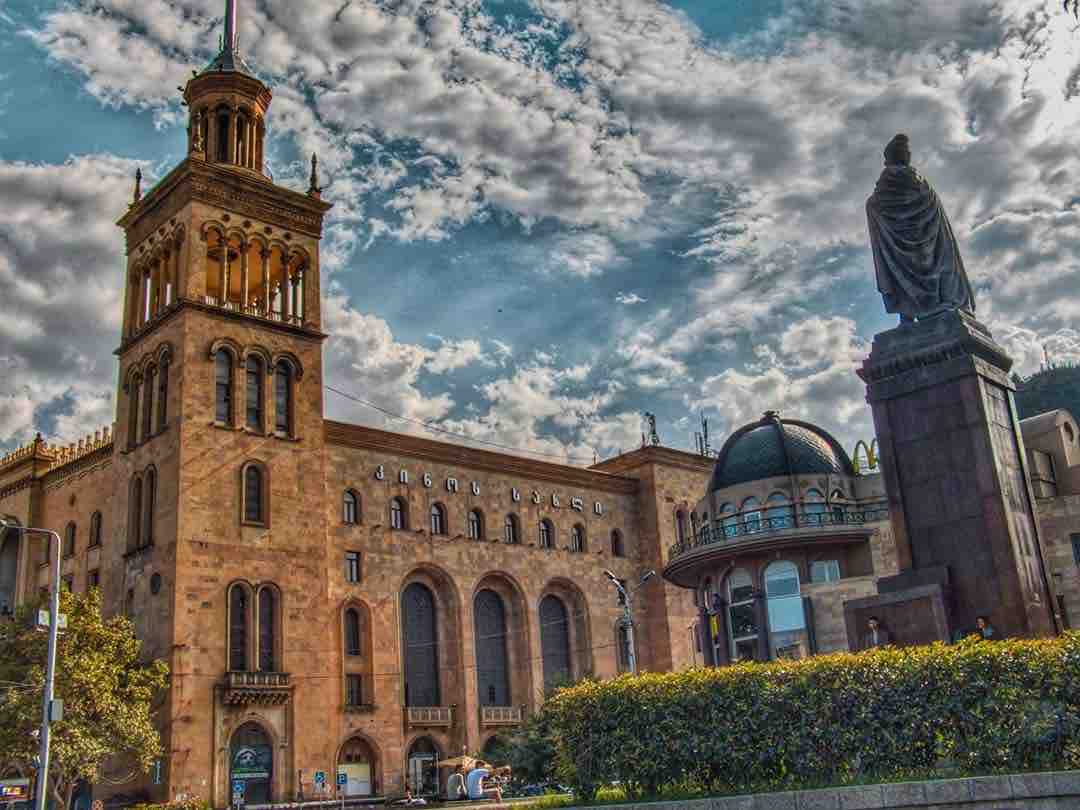 A historical building is featured prominently, showcasing a tall clock tower with detailed architectural elements. A statue stands in the foreground, while lush greenery is visible along the base, and a partly cloudy sky creates a dynamic backdrop.