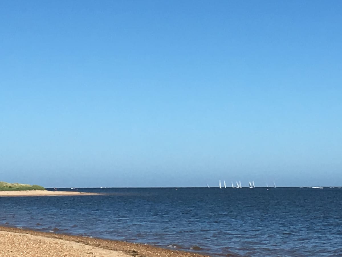 A wide view of the coastline features a pebbled beach fading into calm blue water. The horizon displays several sailboats gliding across the sea under a clear blue sky, providing a serene maritime atmosphere. Gentle waves lap at the shore, inviting leisurely strolls.