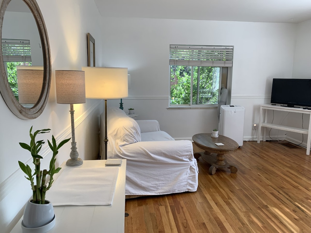 A cozy living space features a white sofa and a round wooden coffee table. A lamp and a decorative plant are positioned on a side table. Light filters in through a window with blinds, creating a relaxed atmosphere. A television is set on a white console nearby.
