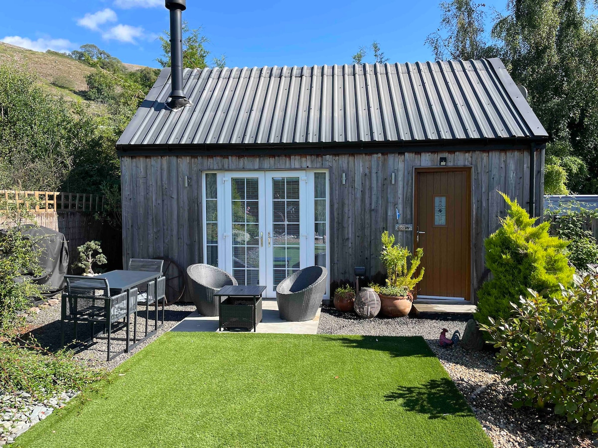 A contemporary wooden structure is showcased with a metal roof, complemented by a neatly manicured lawn. The entrance features a stylish door, while patio furniture offers seating outside. French doors provide an inviting view of the surrounding garden and hills.