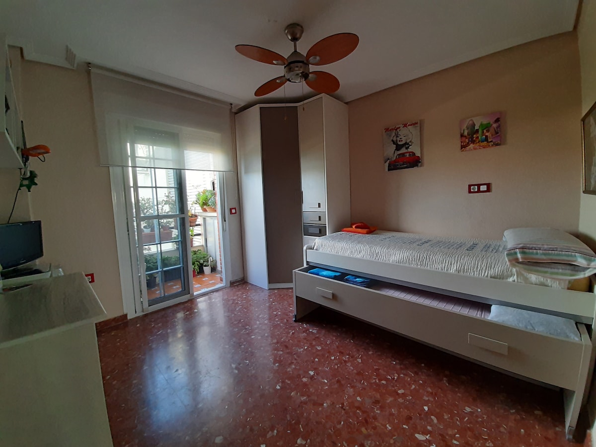 A spacious room is highlighted by a single bed with storage underneath. Natural light filters through a sliding glass door that leads to a small private terrace. A ceiling fan provides a comfortable atmosphere, while wall-mounted storage displays an organized space.