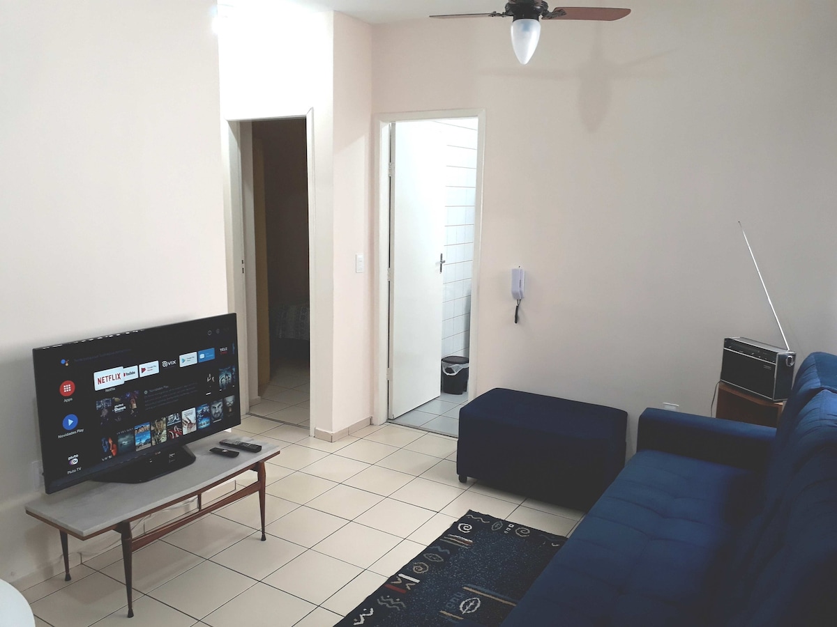 A living room area features a blue sofa and a flat-screen TV with smart features. A small coffee table sits in front of the television. Natural light illuminates the space, highlighting tiled flooring and a ceiling fan.