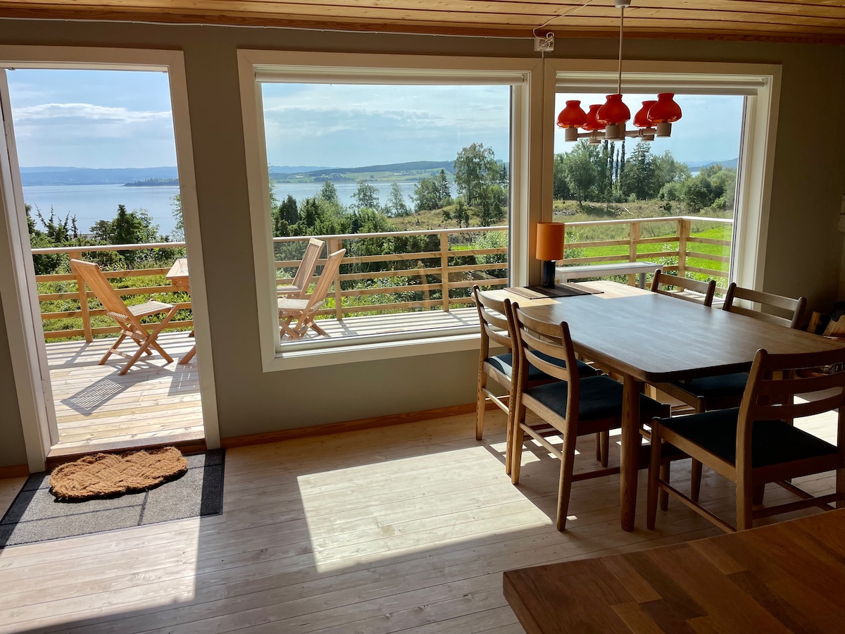 Large windows frame scenic views of the fjord and surrounding landscape. A dining table occupies the center of the room, with wooden chairs arranged around it. Light floods the space, while cozy outdoor seating is visible on the terrace beyond.