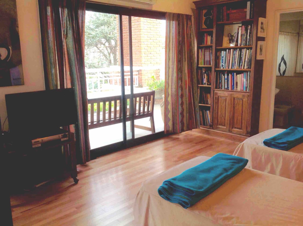 A spacious bedroom features two beds with folded blue towels on top. A wooden bookshelf filled with books is positioned beside a television. Large glass doors open to a private terrace with outdoor seating, allowing natural light to fill the room.