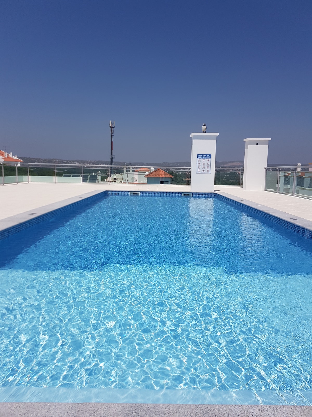 A rooftop saltwater pool is shown, reflecting clear blue skies above. White tiles surround the pool, contributing to a bright and open ambiance. Scenic views of the surrounding landscape are visible in the background, enhancing the tranquil setting.