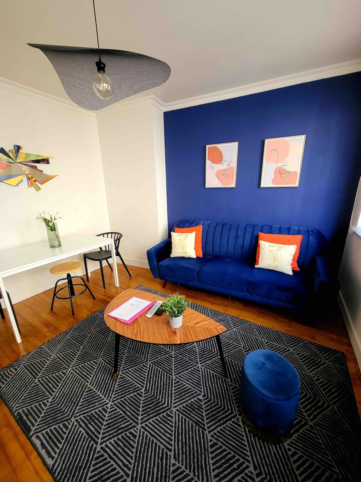 A cozy living area features a blue sofa adorned with accent pillows, positioned next to a round coffee table. A small dining table with chairs is visible, along with a decorative wall piece and two framed artworks. Natural light fills the space through a window.