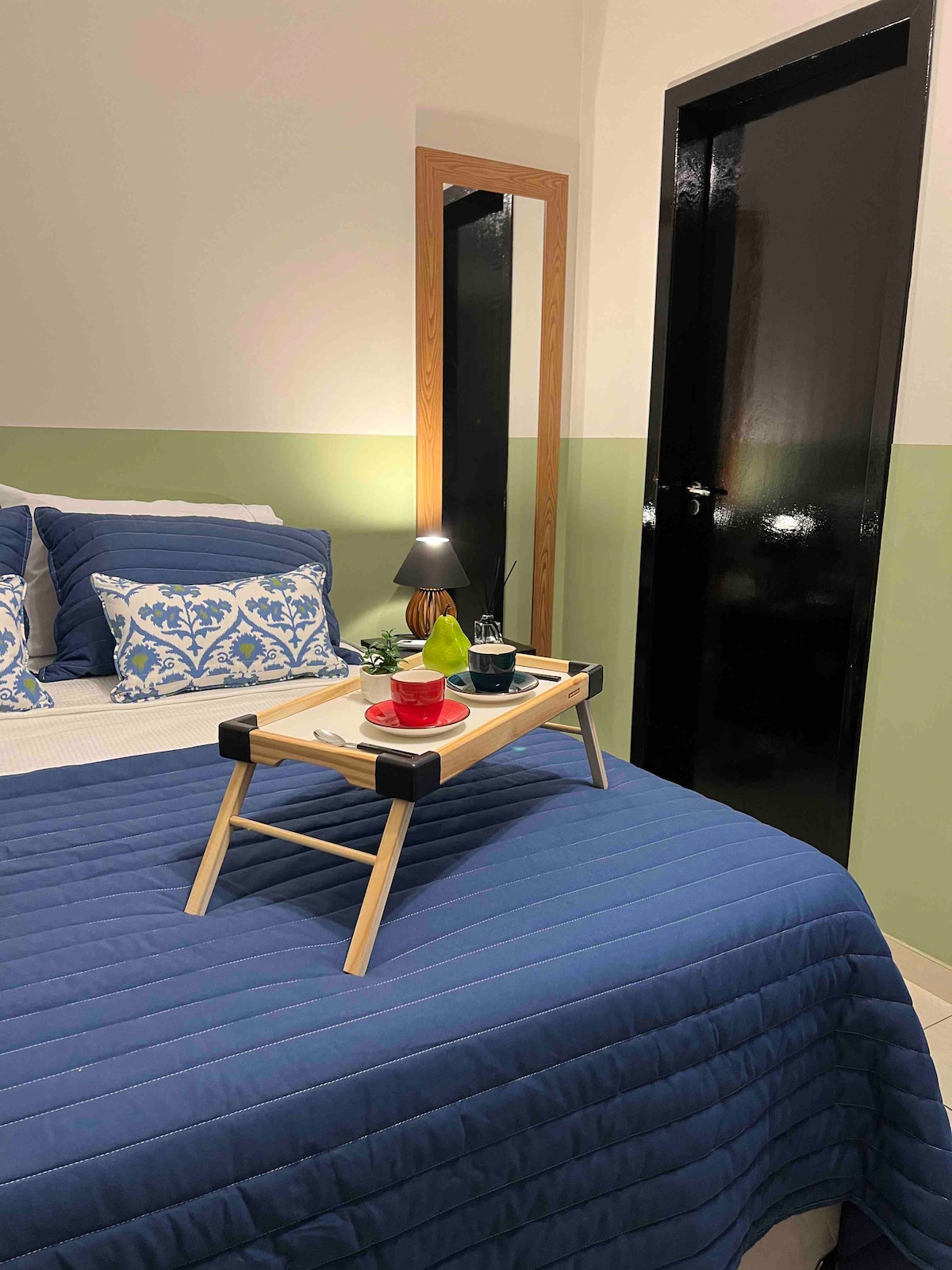A spacious bed is dressed in a deep blue coverlet, topped with embroidered pillows. A small wooden tray holds snacks and drinks, placed in the center. A mirror and dark door are visible in the background, adding depth to the indoor setting.