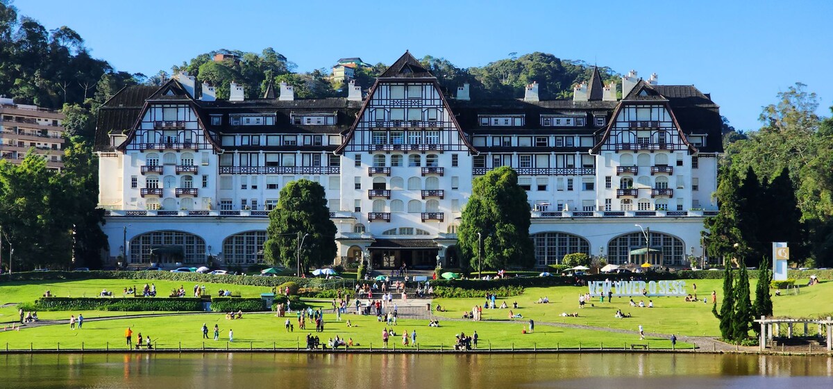 The iconic Quitandinha Hotel is displayed against a clear blue sky, showcasing its distinctive architecture. Lush green lawns extend in front, where visitors can be seen enjoying the outdoor space. The serene lake is visible in the foreground, enhancing the tranquil setting.