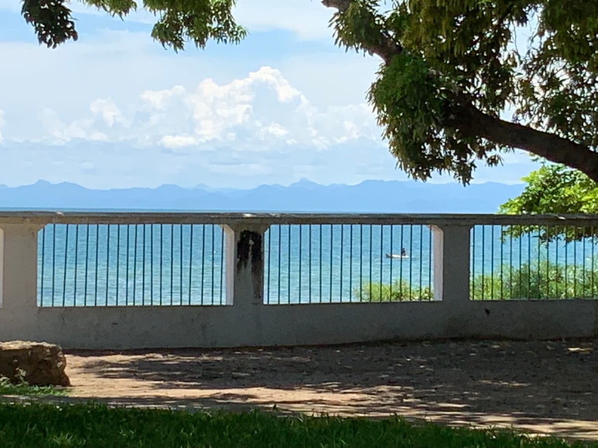 A view of Lake Malawi is captured through a white decorative fence, revealing tranquil waters and a distant fishing boat. Lush greenery frames the scene, with trees providing shade and enhancing the natural serenity of the lakeside setting.