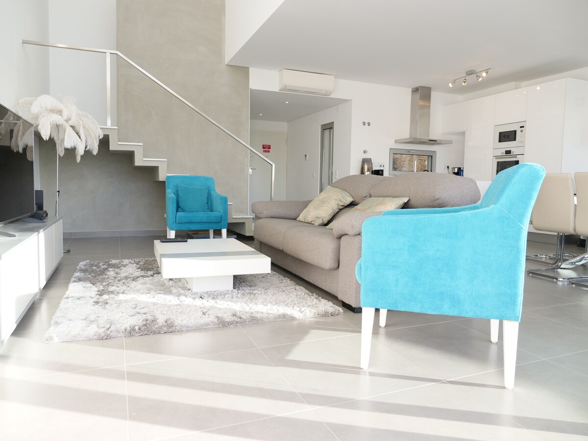 An airy living room is highlighted by a modern gray sofa and two vibrant blue chairs. A sleek coffee table is positioned on a soft area rug. The open layout reveals a contemporary kitchen area and a staircase leading to the upper level.