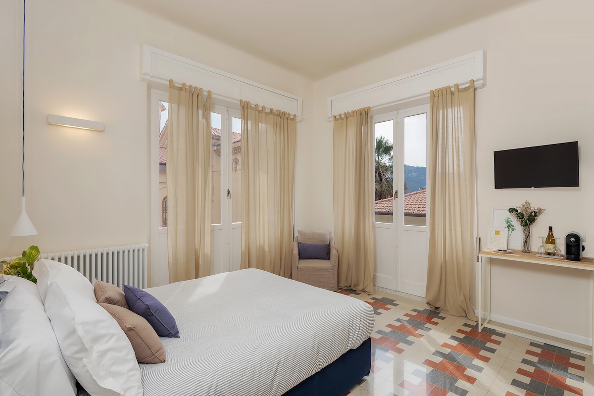 A bright bedroom features a king size bed with layered bedding, complemented by large windows dressed with sheer curtains. A small seating area is provided, alongside a wall-mounted television and a compact table with a coffee maker. The floor showcases a colorful tile design.