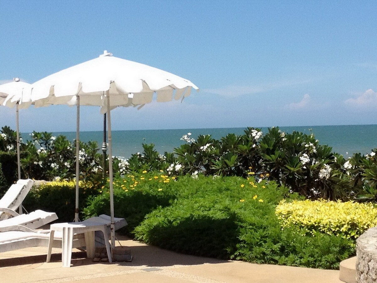 A sunlit outdoor area features a white umbrella offering shade beside a poolside. A pair of lounge chairs rests nearby, surrounded by vibrant green foliage and colorful flowers. The serene ocean view stretches seamlessly into the blue sky in the background.