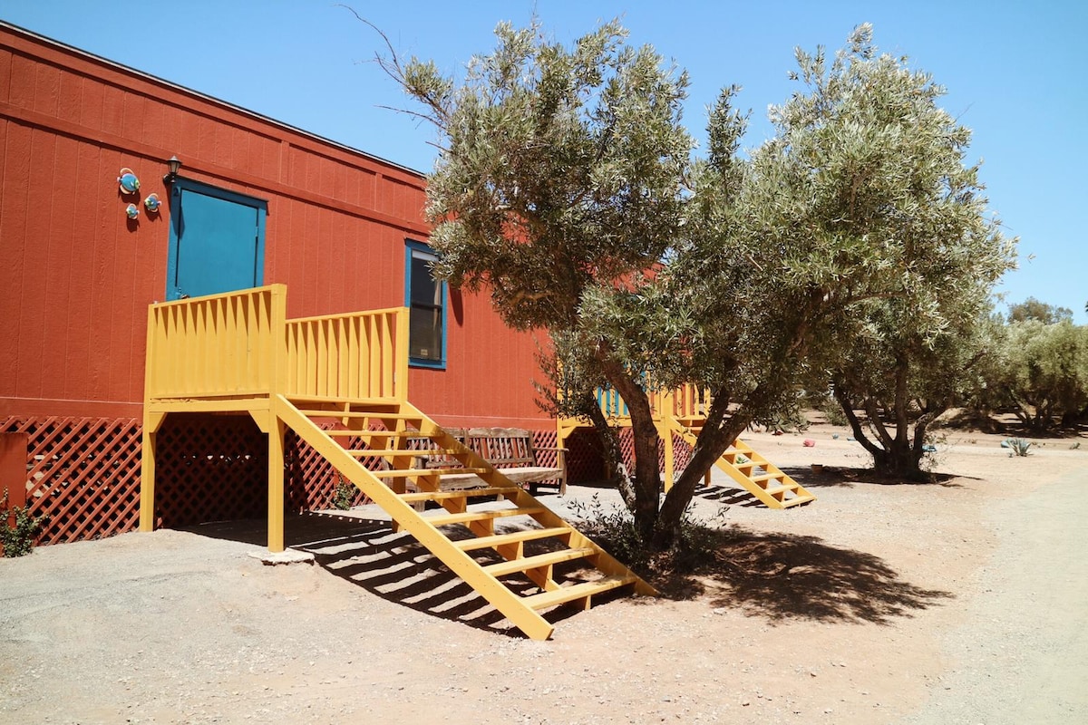 A vibrant exterior of a suite features bold red walls and yellow stairs leading up to a welcoming entrance. Olive trees provide shade around the entrance, enhancing the natural setting. The pathway is simple and clear, allowing for easy access to the suites.