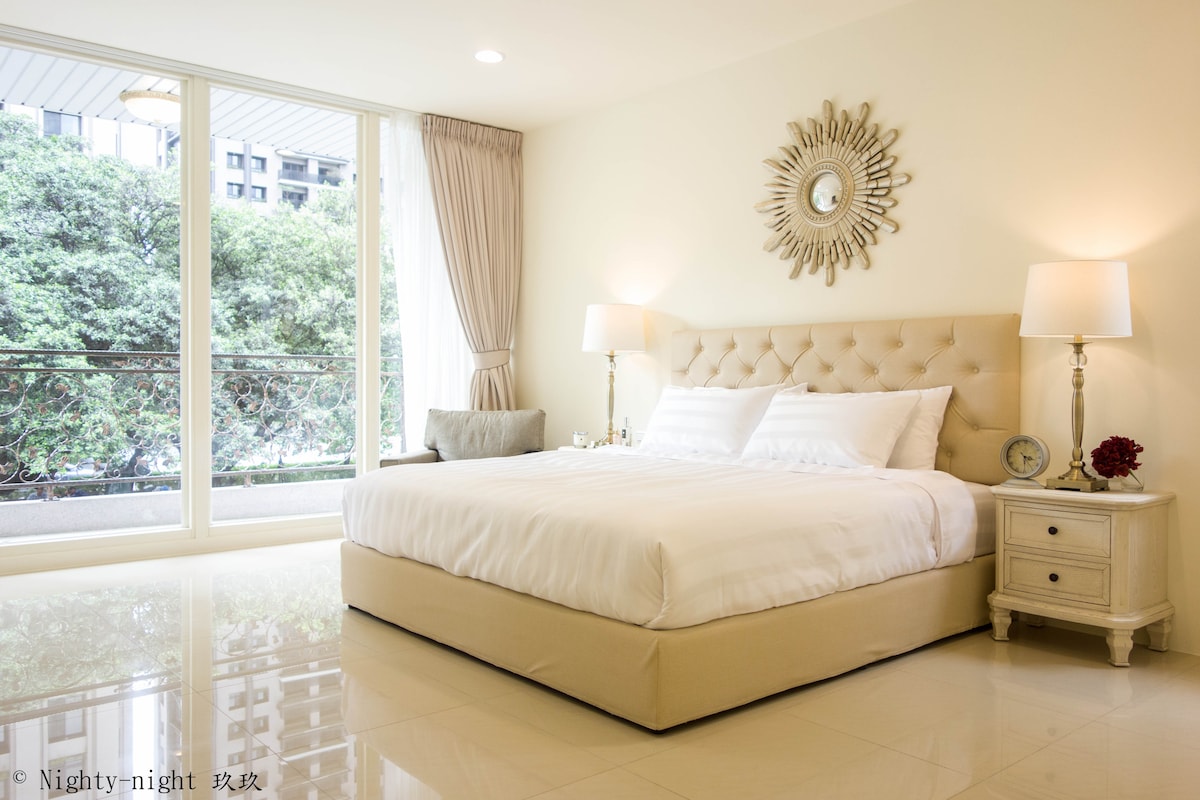 A spacious bedroom features a queen size bed with a tufted beige headboard, flanked by bedside tables with lamps. Large glass doors lead to a balcony, providing ample natural light. A decorative mirror is hung above the bed, enhancing the room's elegance.