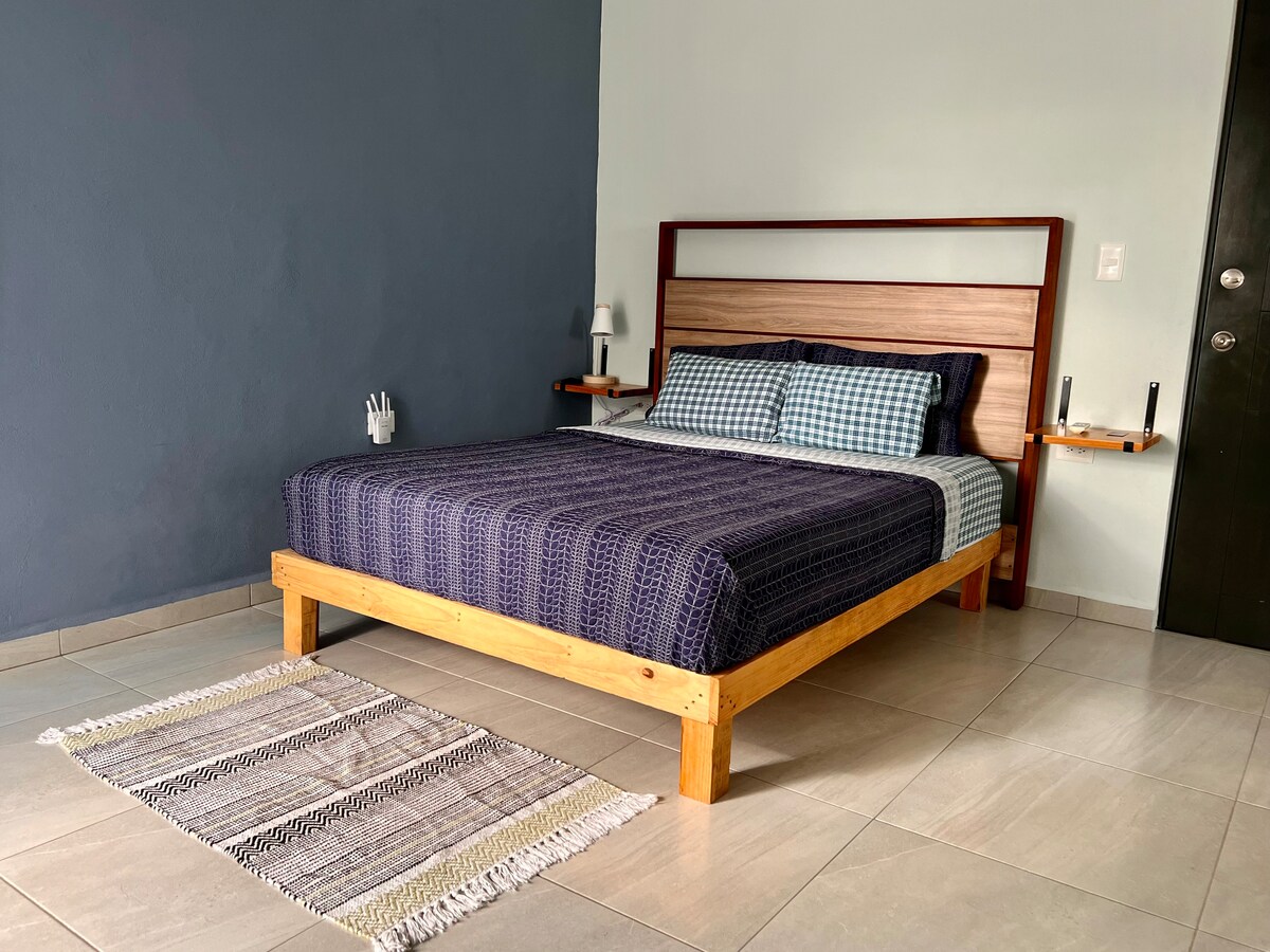 A cozy sleeping area features a wooden bed with a checkered blanket and decorative pillows. Side tables are positioned on either side, each holding a lamp. The room is complemented by a small woven rug on the tiled floor, with a light blue wall providing contrast.