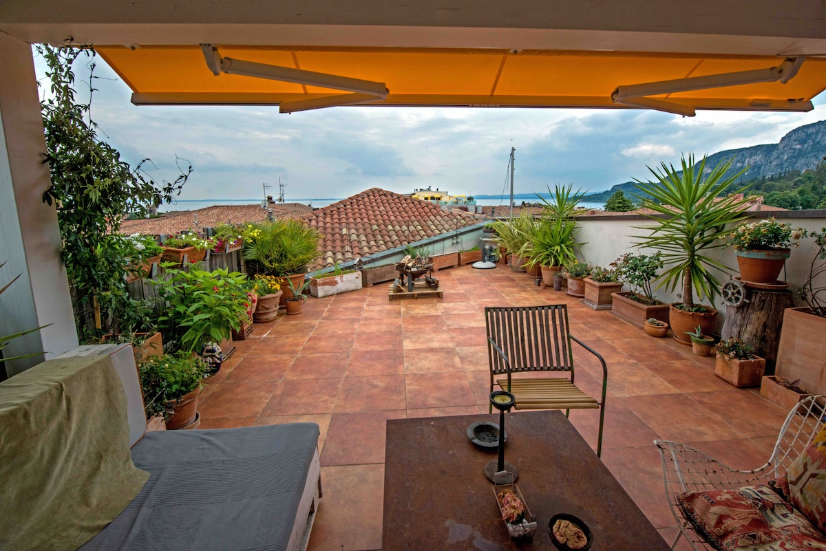 A spacious rooftop terrace is depicted, featuring terracotta tiles and surrounded by an array of potted plants. A small table with bowls sits in the foreground, while a comfortable seating area and an umbrella provide shaded relaxation with views of the sea and rooftops.
