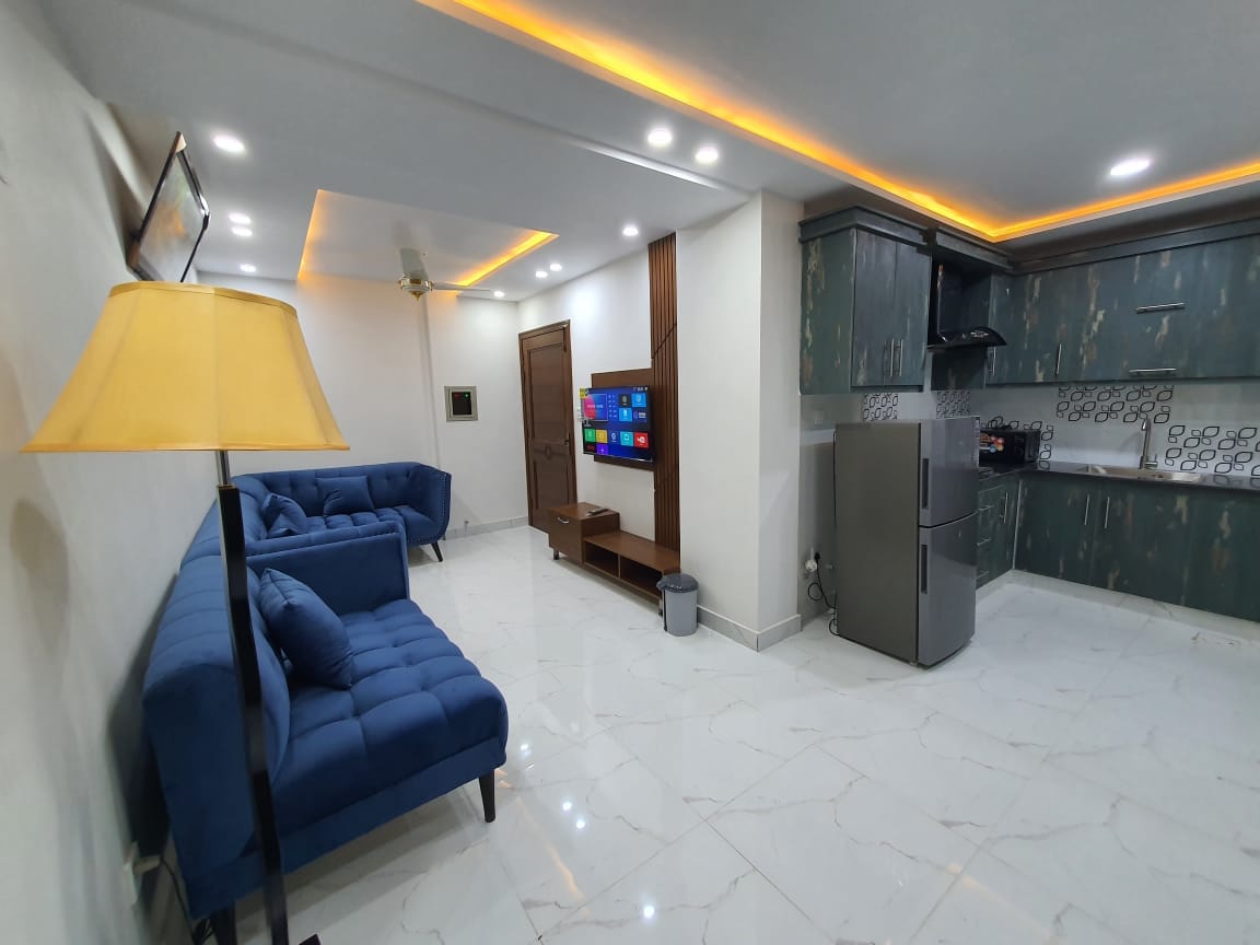 A modern living space features a blue velvet sofa and a sleek entertainment unit with a flat-screen TV. Recessed lighting enhances the ceiling, while a stylish lamp provides additional illumination. The open kitchen is equipped with contemporary cabinetry and appliances, creating a functional yet elegant atmosphere.