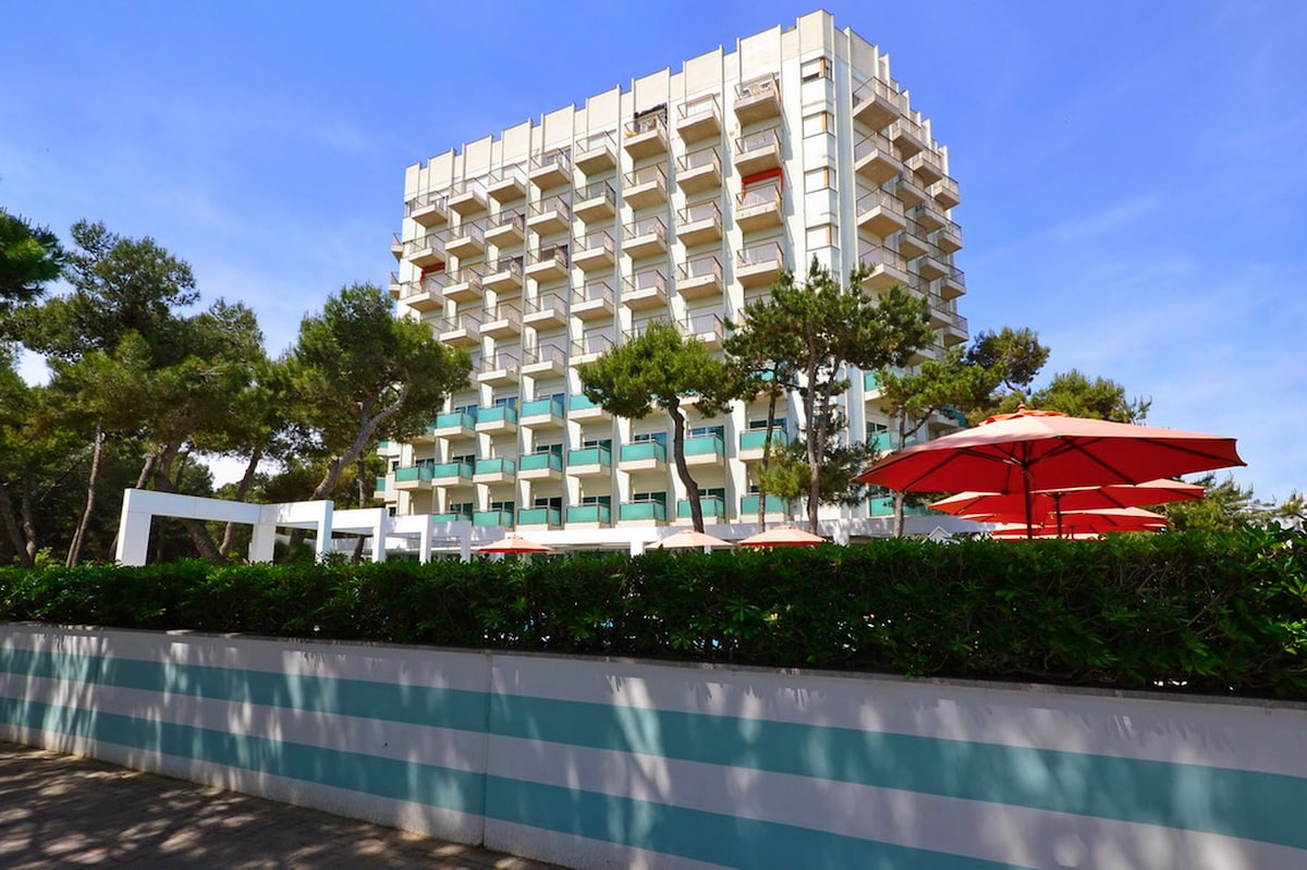 A multi-story building is visible, featuring balconies and large windows. Surrounding greenery includes trees and bushes, providing a tranquil environment. Red umbrellas are set up for outdoor seating, contributing to the overall welcoming atmosphere near the entrance.