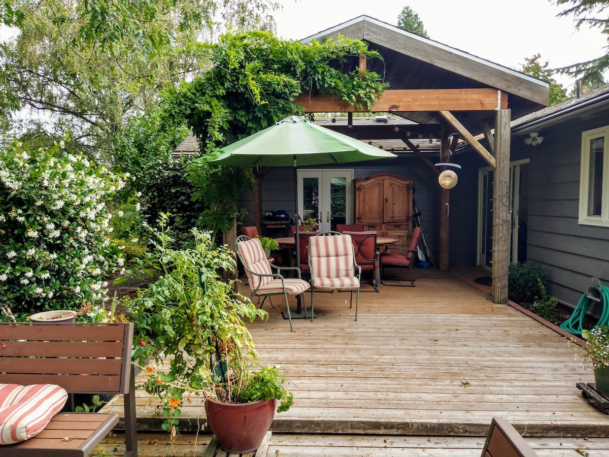 A wooden deck features a shaded seating area with striped chairs and a large green umbrella. Lush greenery surrounds the space, including flowering plants and trellises. A dining table is positioned nearby, providing a suitable setting for outdoor meals and relaxation.