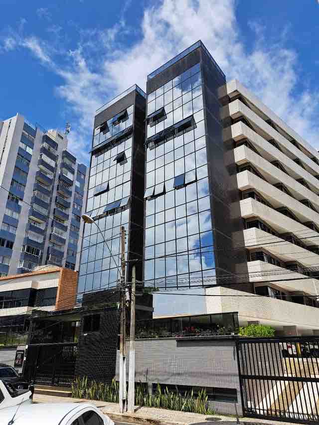 A modern building with a sleek glass façade is displayed against a blue sky with scattered clouds. The structure features multiple stories with a combination of reflective glass and solid surfaces, showcasing an entrance with a security gate surrounded by greenery.