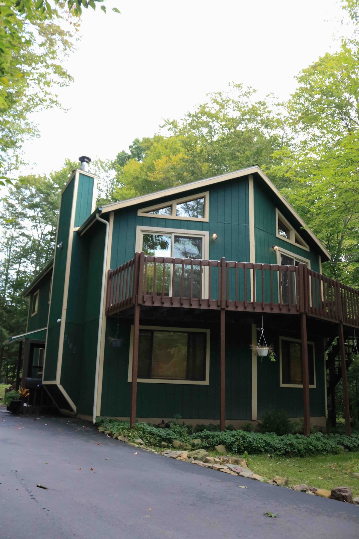 The exterior of a green-painted house, surrounded by trees, showcases a two-story structure with large windows. A balcony is visible above the entrance, and a paved driveway leads to the front. Natural landscaping features stones and shrubs, contributing to the tranquil setting.