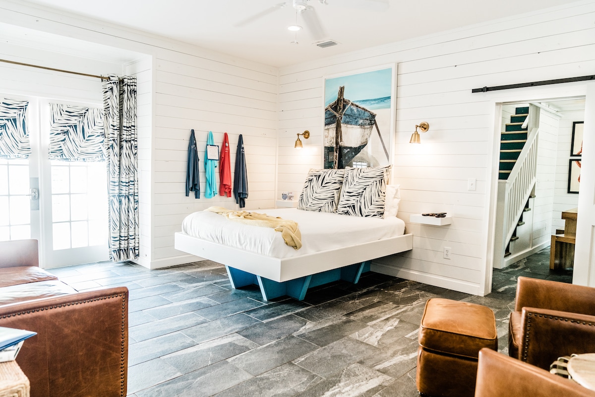 A creatively designed studio features a signature floating bed with vibrant zebra-patterned bedding. Large windows provide natural light and an open view to the outdoor area. Comfortable leather club chairs complement the space, while beach-inspired artwork adds a touch of character.