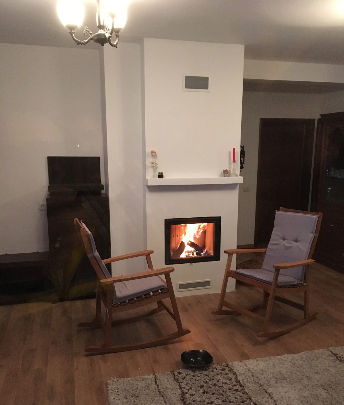 Two rocking chairs are positioned near a modern fireplace, featuring a warm flame. A light-colored area rug lies on the wooden floor, enhancing the cozy atmosphere. A wall-mounted vent and nearby storage cabinet are visible, contributing to the inviting interior space.