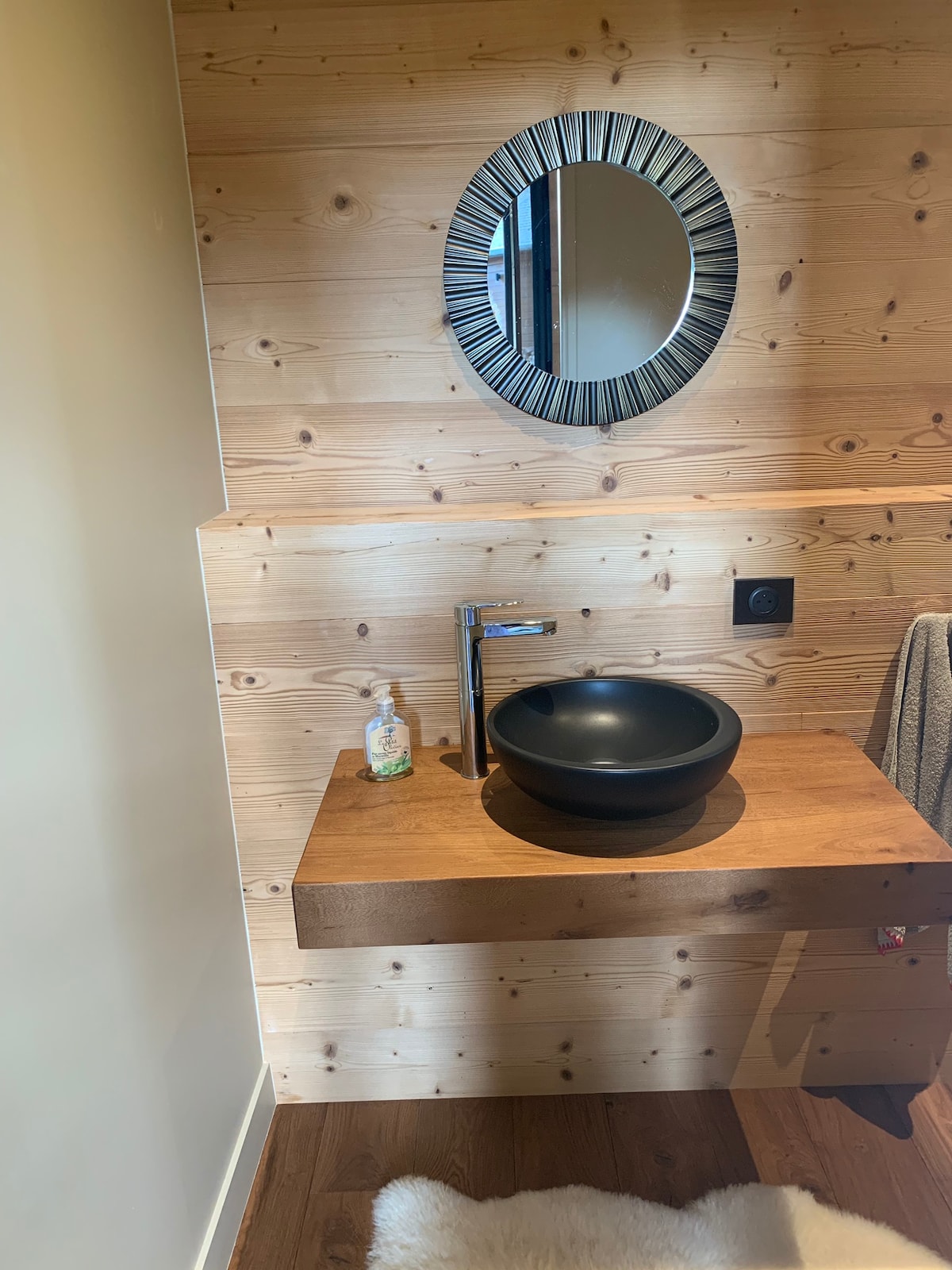 A modern bathroom space features a wooden wall adorned with natural textures. A circular mirror reflects the room's light above a wooden countertop, which holds a round black basin and a soap dispenser. A towel hangs nearby, enhancing the overall functionality.