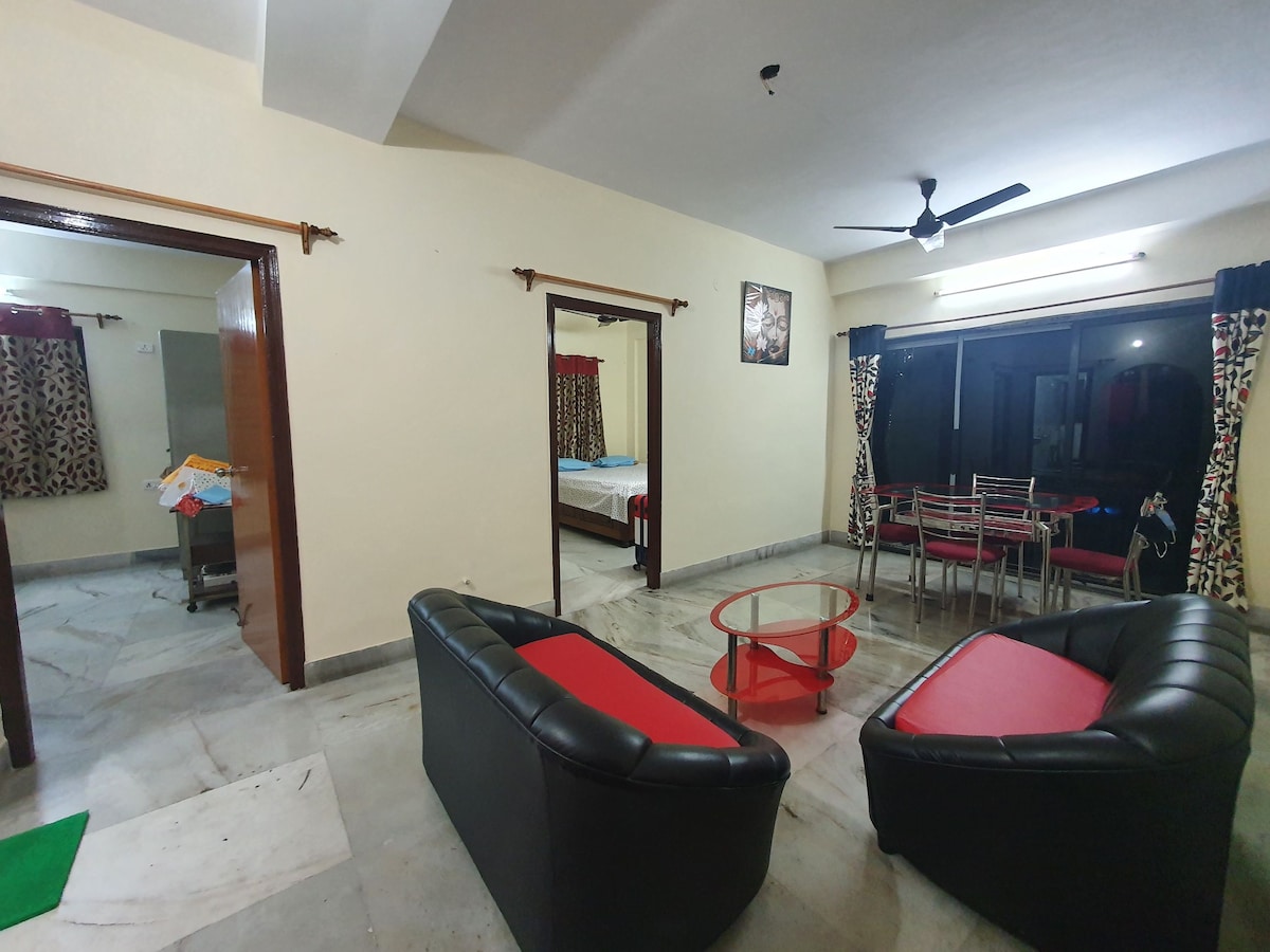 A spacious living area is presented, featuring a pair of black and red sofas surrounding a circular table. A modern dining area with four chairs is visible by a large window, allowing natural light. Two doorways lead to the bedrooms, enhancing the open feel.
