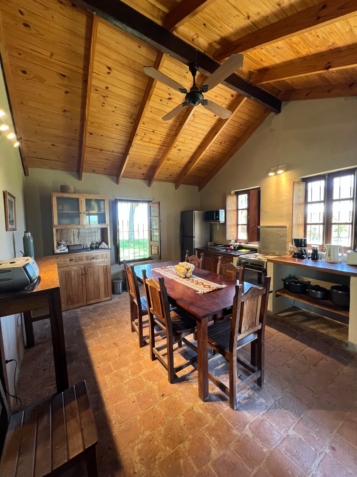 The rustic kitchen features a wooden dining table surrounded by chairs. High ceilings with exposed beams add to the spacious feel. A dark stove and various kitchenware are visible on the counters. Natural light enters through multiple windows, illuminating the warm tones of the decor.