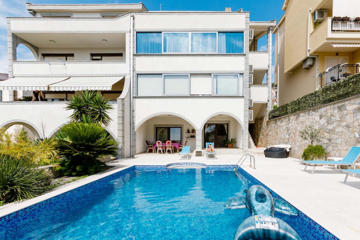 A modern villa is framed by a sparkling blue swimming pool, reflecting the clear sky above. The pool area features lounge chairs, and colorful seating is visible on a shaded terrace. Lush greenery surrounds the building, enhancing the tranquil environment.