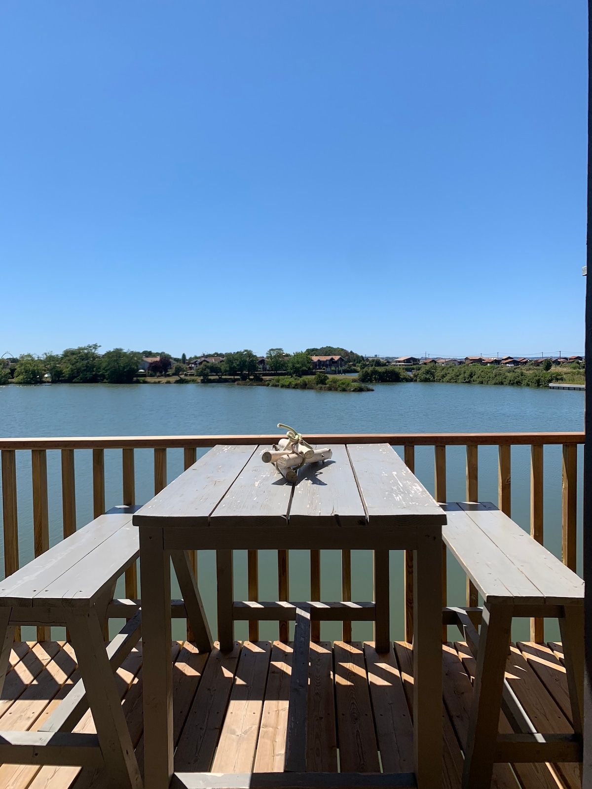 A wooden terrace features a rectangular table accompanied by two stools, overlooking a calm water body. The clear blue sky complements the serene setting, while greenery is visible along the shore, providing a peaceful backdrop.