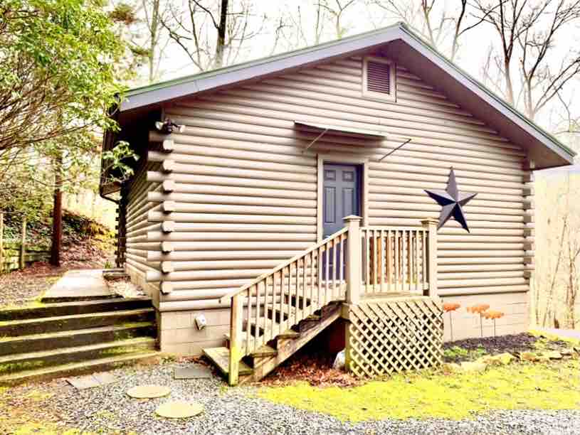 The exterior of a log cabin features a sturdy wooden staircase leading to the front door, framed by a small porch. Dark wooden accents, including a star decoration, are present on the cabin's side. The surrounding greenery enhances the secluded mountain setting.