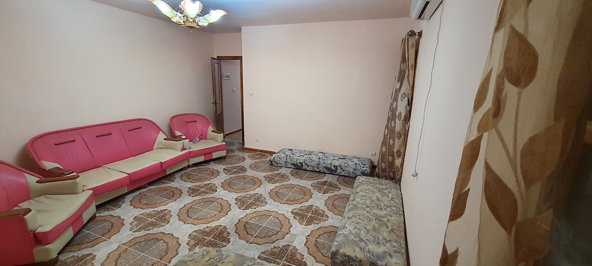 A spacious living area features pink upholstered couches arranged around a patterned tile floor. Soft seating options are provided, including cushions and a large rug. Natural light gently illuminates the room, complemented by soft curtains and a decorative overhead light fixture.