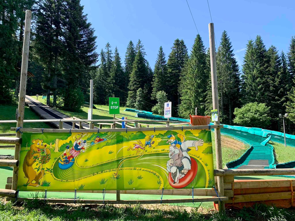 A vibrant mural showcases colorful cartoon characters engaging in fun activities on a winding track. The backdrop features lush green trees under a clear blue sky, complementing the lively scene that emphasizes outdoor enjoyment at the recreational area.