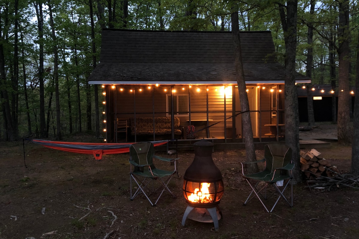 A cabin exterior is illuminated by string lights, surrounded by trees. A fire pit with flames can be seen in the foreground, accompanied by two camping chairs. A hammock is strung nearby, creating a tranquil outdoor space for relaxation.